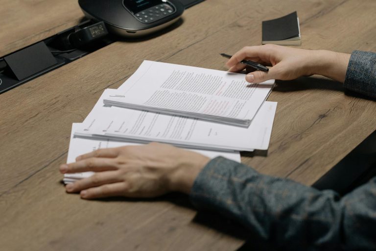 Close-up of a person reviewing documents with a pen at a modern office desk.